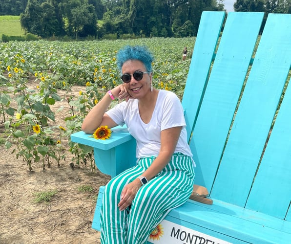 Sharman Monroe smiling and leaning in a giant turquoise chair in a sunflower field, symbolizing joyful freedom, personal empowerment, and living life on your own terms